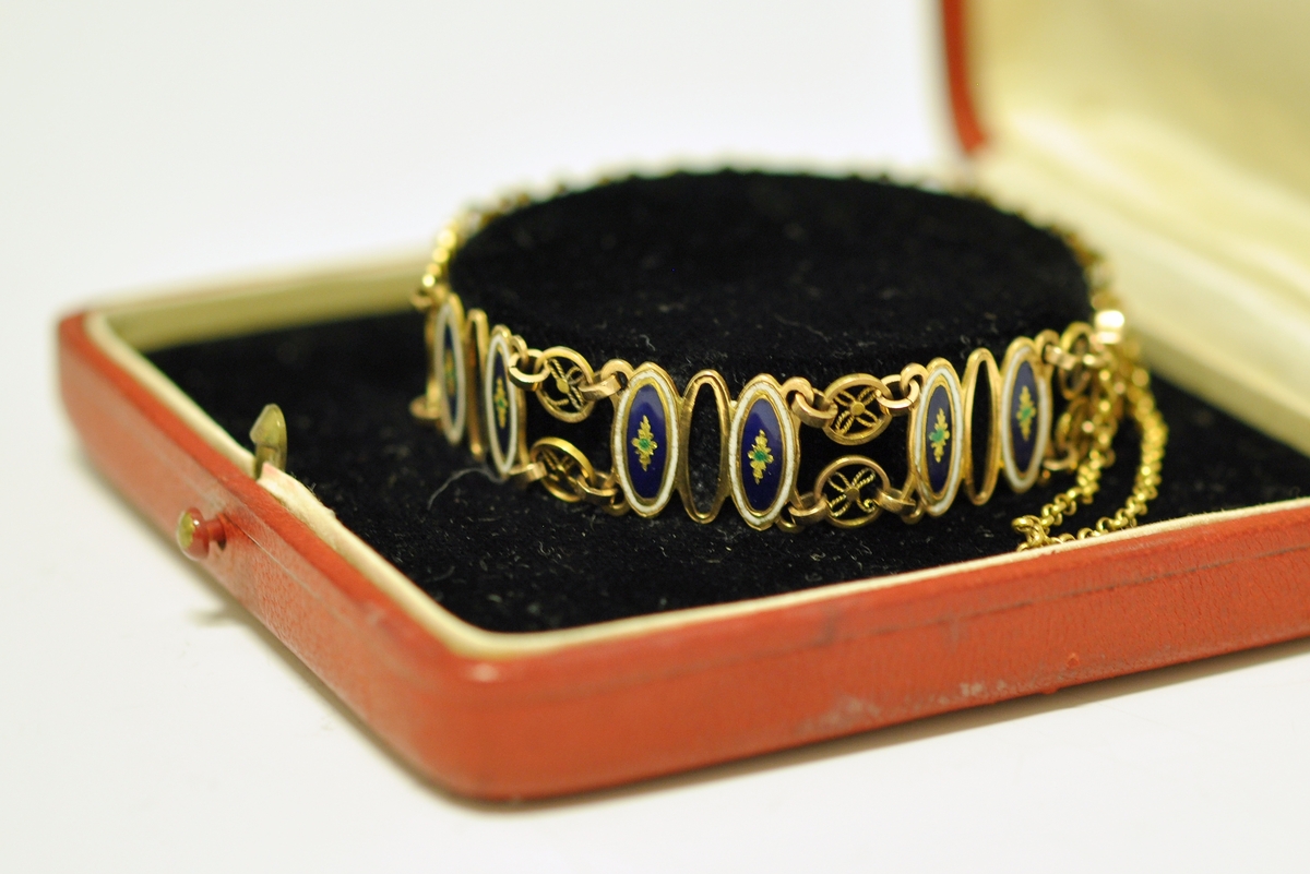 Armbånd av gull og emalje. Sammenleddet, hvert ledd består av tre ovale former, det midterste åpent, de to andre emaljert. De emaljerte feltene har hvit emalje ytterst og blå innerst med en stjerneformet blomst i gult og grønt i midten. Hjulformer binder leddene sammen. Låsen rektangulær med butte hjørner og svakt innsvingede sider. Sisselert med blomst. Tynn lenke mellom lås og spenne. Ustemplet. Fra slutten av 1800 tallet.