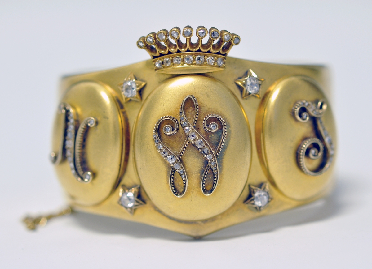 A.Bredt gullarmbånd som kan åpnes  på siden med lås og sikkerhetslenke. Armbåndet går i en spiss foran og det er montert en krone øverst men flere små diamanter, kronen er noe løs. Foran har armbåndet tre lokk  som kan åpnes. Framsiden av lokkene er dekoret med initialer i diamanter :  NWJ. Hvert lokk kan åpnes og inneholder fire fotografier av mannspersoner.Innvendig er det gravert inn årstall og stemplet.

B. Lilla eske smykkeeske som er tilpasset armbåndets form og har lås. Innvendig i lokket er firmaets navn stemplet.