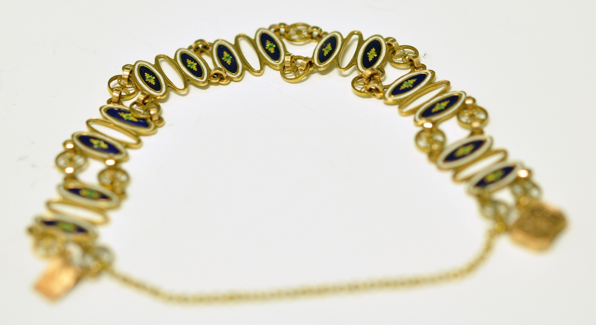 Armbånd av gull og emalje. Sammenleddet, hvert ledd består av tre ovale former, det midterste åpent, de to andre emaljert. De emaljerte feltene har hvit emalje ytterst og blå innerst med en stjerneformet blomst i gult og grønt i midten. Hjulformer binder leddene sammen. Låsen rektangulær med butte hjørner og svakt innsvingede sider. Sisselert med blomst. Tynn lenke mellom lås og spenne. Ustemplet. Fra slutten av 1800 tallet.