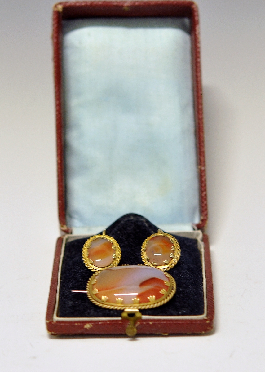 Smykkesett som består av 1 stk brosje, og 2 stk øreheng. A.Oval nesten kvadratisk brosje som består av  brun-orange stein (agatl?). Steinen er omkranset av smal metallkant  som er snodd rundt steinen, og  festet med små "blad" Steinen vises også på baksiden, og en smal nål er montert på.

B. og C. Øreheng/pynt i samme utførelse som brosjen, det er montert en lang bøyle til feste.  

D. Brunrød smykkeeske medfølger, på baksiden kan det være et stempel eller nr?