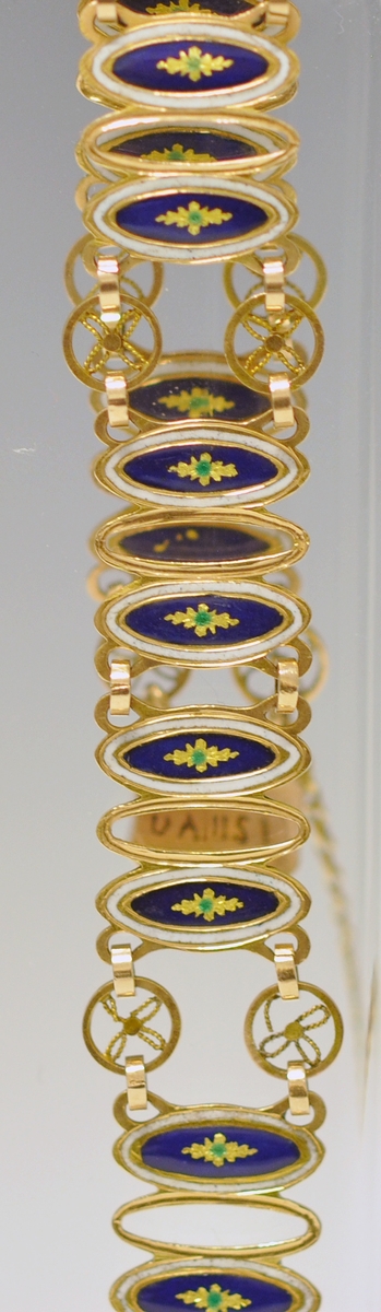 Armbånd av gull og emalje. Sammenleddet, hvert ledd består av tre ovale former, det midterste åpent, de to andre emaljert. De emaljerte feltene har hvit emalje ytterst og blå innerst med en stjerneformet blomst i gult og grønt i midten. Hjulformer binder leddene sammen. Låsen rektangulær med butte hjørner og svakt innsvingede sider. Sisselert med blomst. Tynn lenke mellom lås og spenne. Ustemplet. Fra slutten av 1800 tallet.