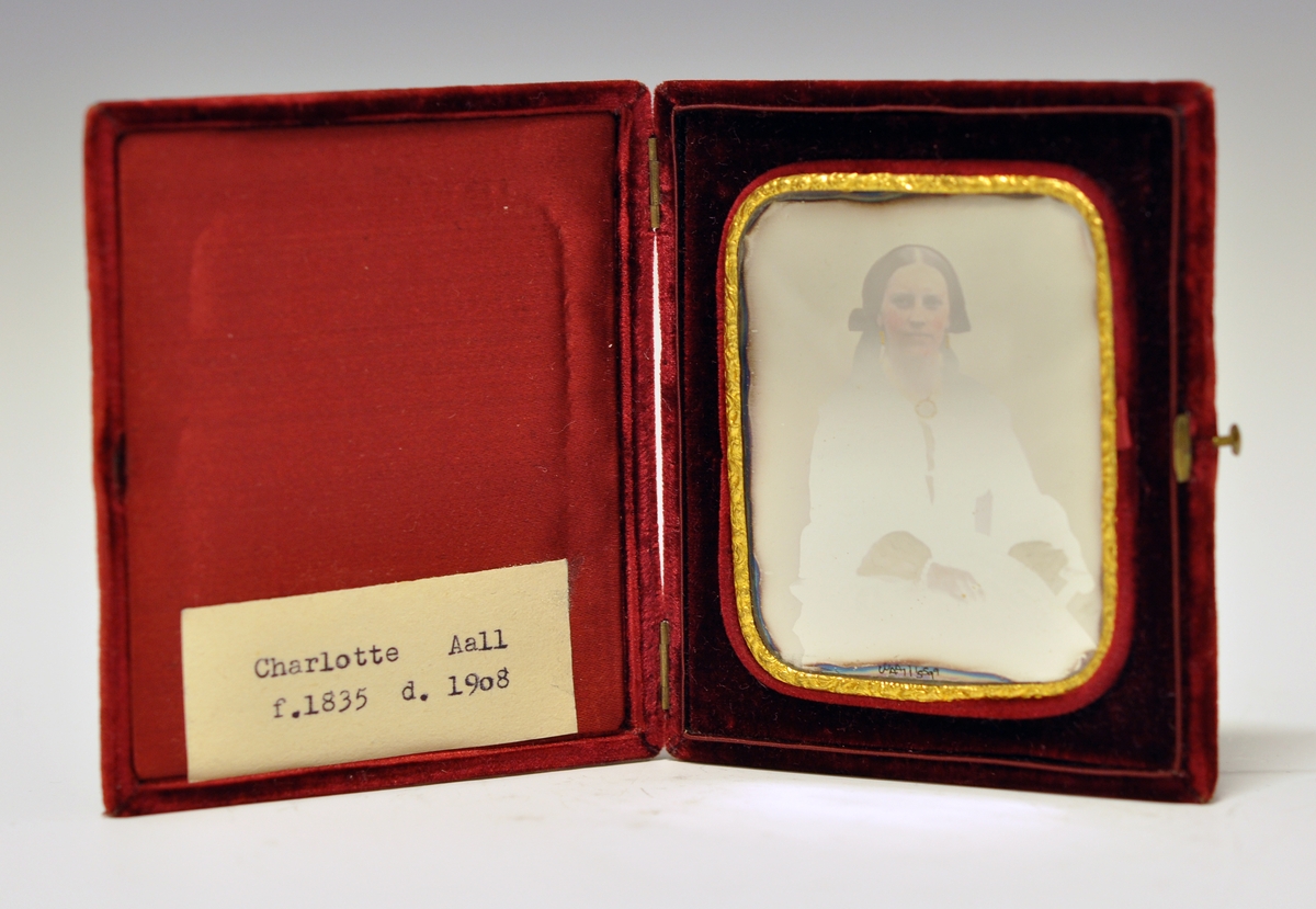 Daguerrotypi i etui. Etui dekket med rød fløyel. To små hengsler og knapp til å åpne. Innvendig portrett av sittende kvinne, gullkant med reileffdekor rundt fotogarfiet, avrundede hjørner. Bildet kan taes ut av etuiet. Kvinnen er Charlotte Aall (1835-1908).