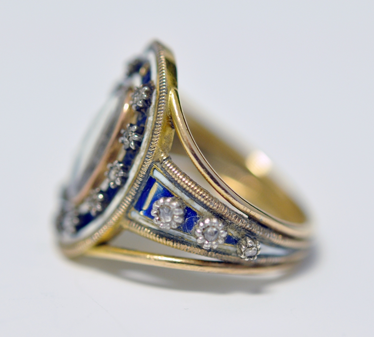 Ring i gull med brillianter, glass, perler og emalje. Tre bånd i gull danner ringen, hvorav de to ytterste er smalest og skiller lag inn mot oval topplate. Den er omkranset av smal gullkant, emaljert flate med stjerneformede brillianter, oval glassflate i midten og med A i perler. Tre brillianter på hver side, på hvitt/blått emaljert felt. Ytterst på emaljert felt og rundt ovalt toppstykke; smalt gullbånd med striper på tvers. Ustemplet.
1700 tallet.