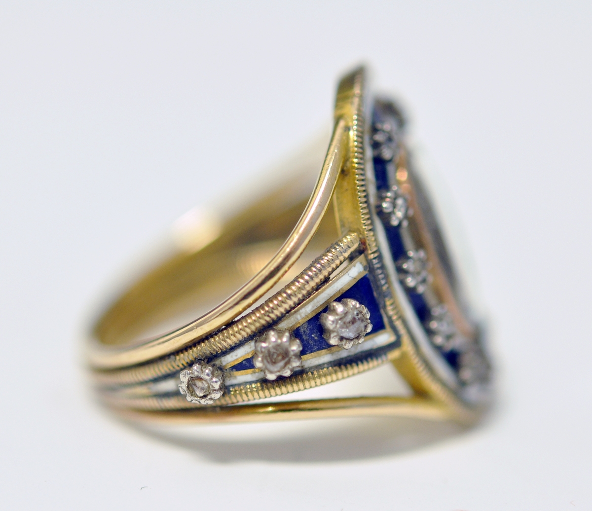 Ring i gull med brillianter, glass, perler og emalje. Tre bånd i gull danner ringen, hvorav de to ytterste er smalest og skiller lag inn mot oval topplate. Den er omkranset av smal gullkant, emaljert flate med stjerneformede brillianter, oval glassflate i midten og med A i perler. Tre brillianter på hver side, på hvitt/blått emaljert felt. Ytterst på emaljert felt og rundt ovalt toppstykke; smalt gullbånd med striper på tvers. Ustemplet.
1700 tallet.