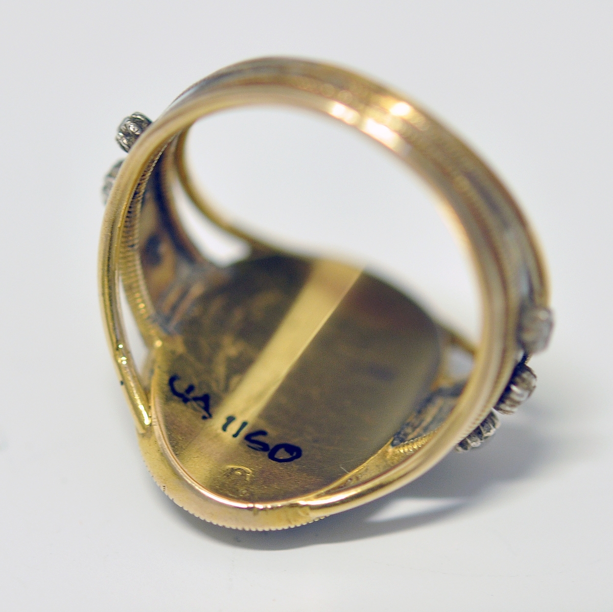 Ring i gull med brillianter, glass, perler og emalje. Tre bånd i gull danner ringen, hvorav de to ytterste er smalest og skiller lag inn mot oval topplate. Den er omkranset av smal gullkant, emaljert flate med stjerneformede brillianter, oval glassflate i midten og med A i perler. Tre brillianter på hver side, på hvitt/blått emaljert felt. Ytterst på emaljert felt og rundt ovalt toppstykke; smalt gullbånd med striper på tvers. Ustemplet.
1700 tallet.