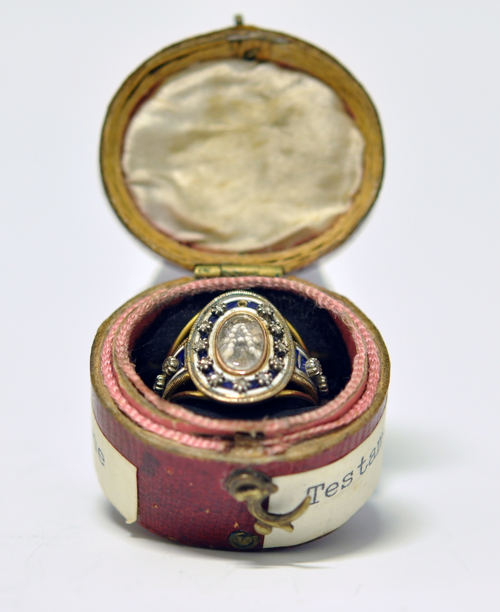 Ring i gull med brillianter, glass, perler og emalje. Tre bånd i gull danner ringen, hvorav de to ytterste er smalest og skiller lag inn mot oval topplate. Den er omkranset av smal gullkant, emaljert flate med stjerneformede brillianter, oval glassflate i midten og med A i perler. Tre brillianter på hver side, på hvitt/blått emaljert felt. Ytterst på emaljert felt og rundt ovalt toppstykke; smalt gullbånd med striper på tvers. Ustemplet.
1700 tallet.