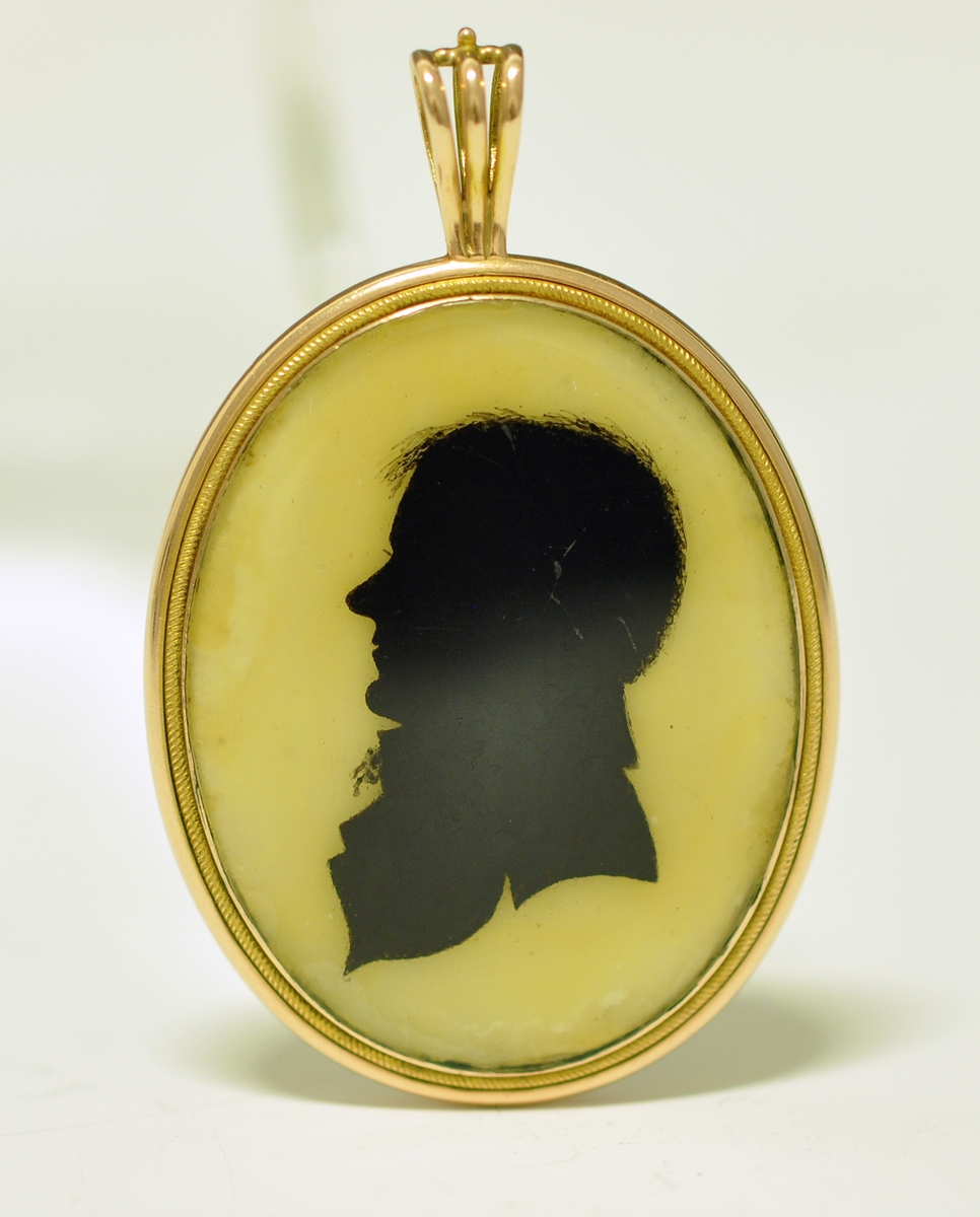 A.Oval medaljong med mann i svart siluett  på gul bunn på fremsiden. Baksiden viser en hårlokk med perler på brun bunn. Begge sider har glass over flatene. Rundt medaljongen er det metall og en dråpeformet metallring delt i tre deler til å tre et kjede inni. Kjedet mangler.
B. Brun smykkeeske tilpasset formen til medaljongen, med lokk og lås. Mørk lilla innvendig med stempel.