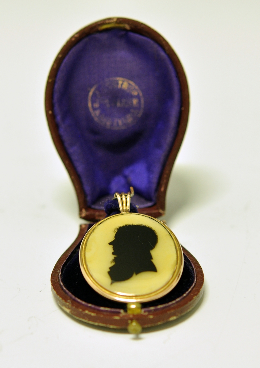 A.Oval medaljong med mann i svart siluett  på gul bunn på fremsiden. Baksiden viser en hårlokk med perler på brun bunn. Begge sider har glass over flatene. Rundt medaljongen er det metall og en dråpeformet metallring delt i tre deler til å tre et kjede inni. Kjedet mangler.
B. Brun smykkeeske tilpasset formen til medaljongen, med lokk og lås. Mørk lilla innvendig med stempel.