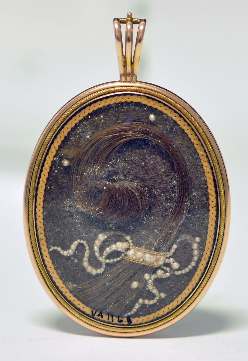 A.Oval medaljong med mann i svart siluett  på gul bunn på fremsiden. Baksiden viser en hårlokk med perler på brun bunn. Begge sider har glass over flatene. Rundt medaljongen er det metall og en dråpeformet metallring delt i tre deler til å tre et kjede inni. Kjedet mangler.
B. Brun smykkeeske tilpasset formen til medaljongen, med lokk og lås. Mørk lilla innvendig med stempel.