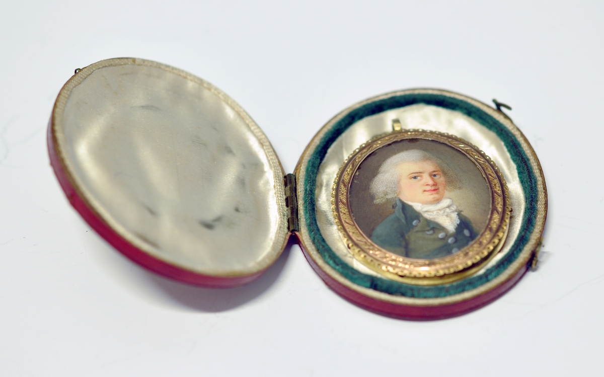 A..Ovalt miniatyrportrett av mann  i midten. Portrettet er innrammet av en smal bølget
perforert krans ytterst i metall og dekket med glass. Øverst oppe er det montert en ring for kjede. Kjedet mangler. Baksiden består av metall. 

B. Oval rød smykkeeske i skinn utvendig og tekstil innvendig.  Har lås og fast lokk som kan åpnes. Ingen stempel.