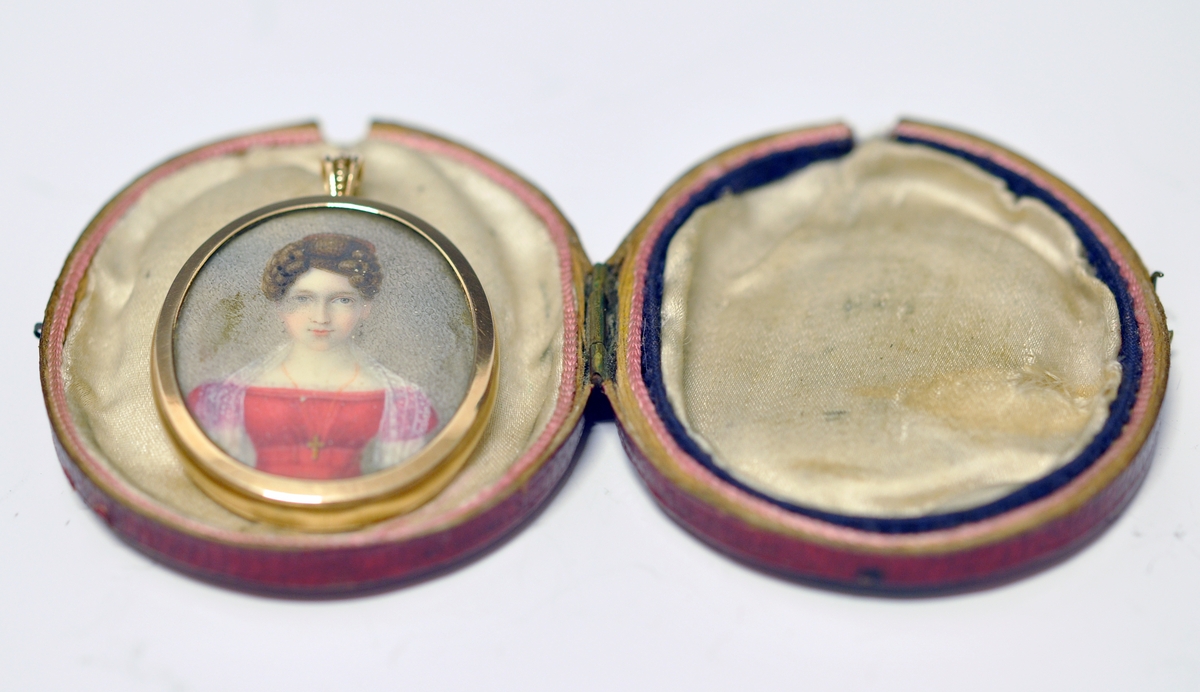 A..Ovalt miniatyrportrett av kvinne  i rød kjole. Portrettet er innrammet av en smal kant i metall og dekket med glass. Øverst oppe er det montert en ring for kjede. Kjedet mangler. På baksiden er det montert en hårlokk med glass over.

B. Oval rød smykkeeske i skinn utvendig og tekstil innvendig.  Har lås og fast lokk som kan åpnes. Ingen stempel.
