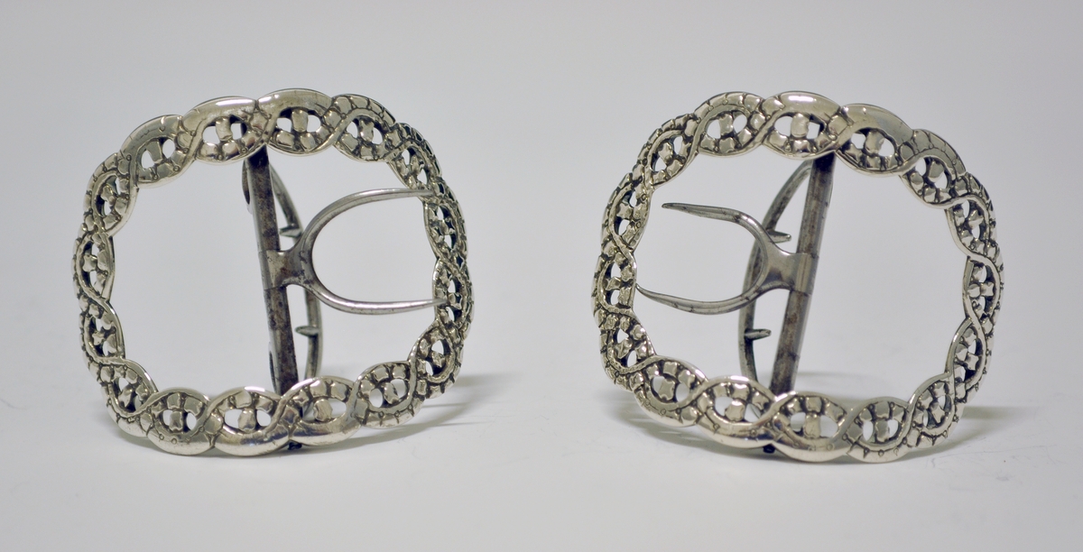Skospenne i sølv. En av et par. Stump, oval form. Flettet, gjennombrutt bånd danner selve spennen. Tverstang med feste til skoen (nyere?).
Stempel: Springende løve. England.
Mester: Samuel Wells, London.