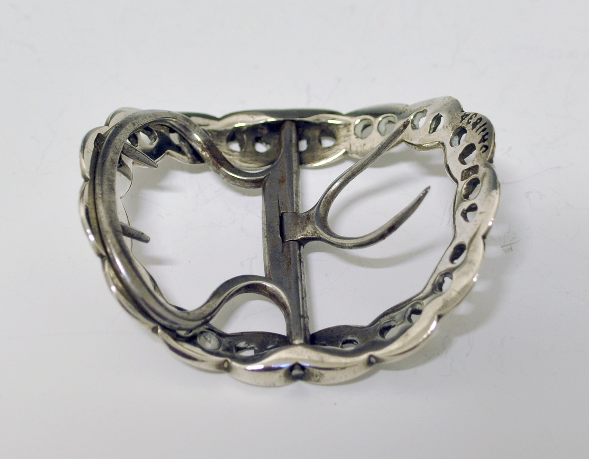 Skospenne i sølv. En av et par. Stump, oval form. Flettet, gjennombrutt bånd danner selve spennen. Tverstang med feste til skoen (nyere?).
Stempel: Springende løve. England.
Mester: Samuel Wells, London.