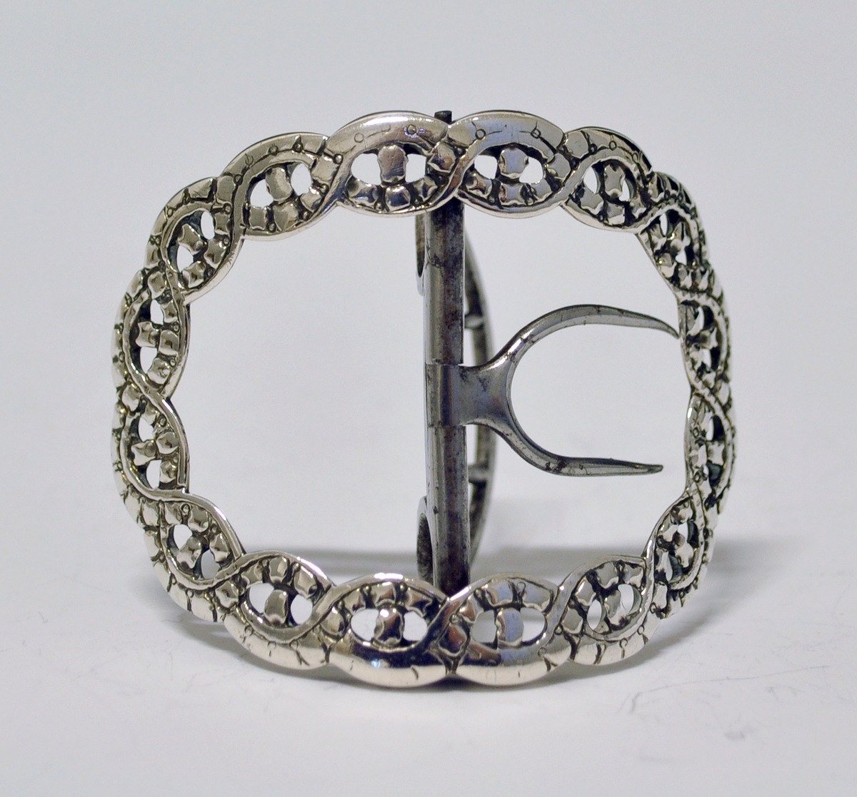 Skospenne i sølv. En av et par. Stump, oval form. Flettet, gjennombrutt bånd danner selve spennen. Tverstang med feste til skoen (nyere?).
Stempel: Springende løve. England.
Mester: Samuel Wells, London.