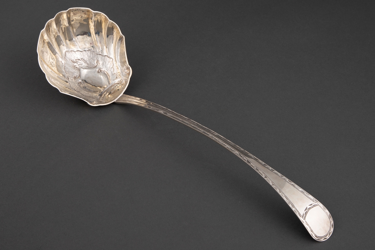 Sølv suppeøse med smalt, lett buet skaft og buet, litt bredere endestykke. Muslingeformet blad. Gravert bord langs skaft på forside og flammeformet gravyr bak på endestykket.