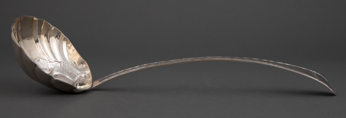 Sølv suppeøse med smalt, lett buet skaft og buet, litt bredere endestykke. Muslingeformet blad. Gravert bord langs skaft på forside og flammeformet gravyr bak på endestykket.