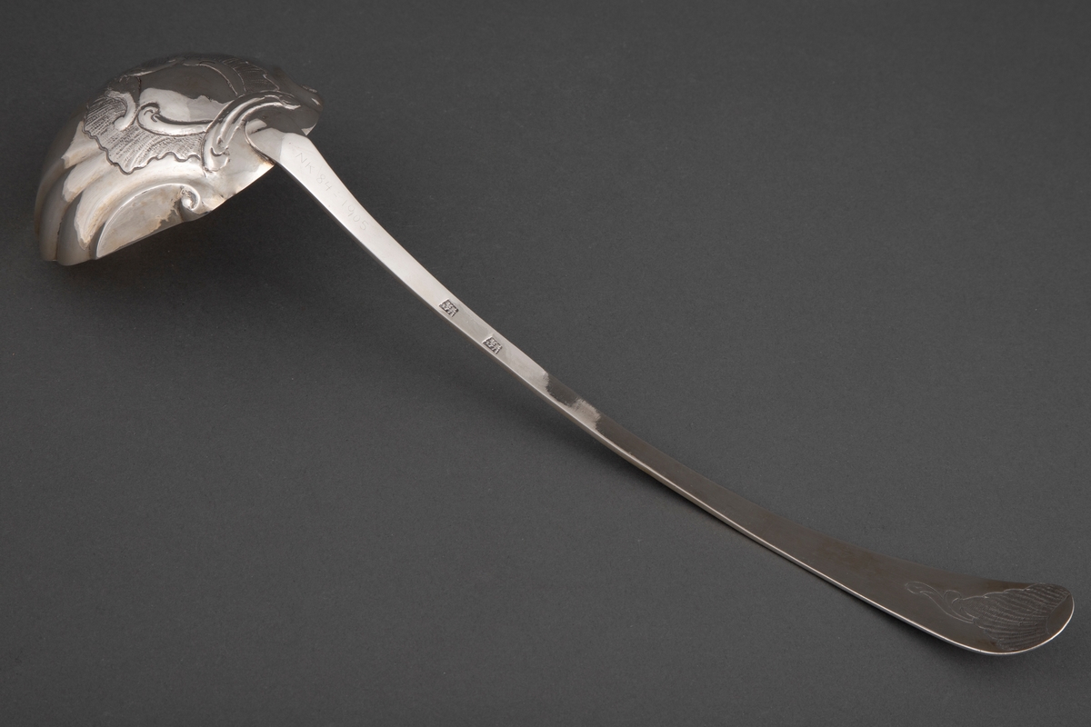 Sølv suppeøse med smalt, lett buet skaft og buet, litt bredere endestykke. Muslingeformet blad. Gravert bord langs skaft på forside og flammeformet gravyr bak på endestykket.