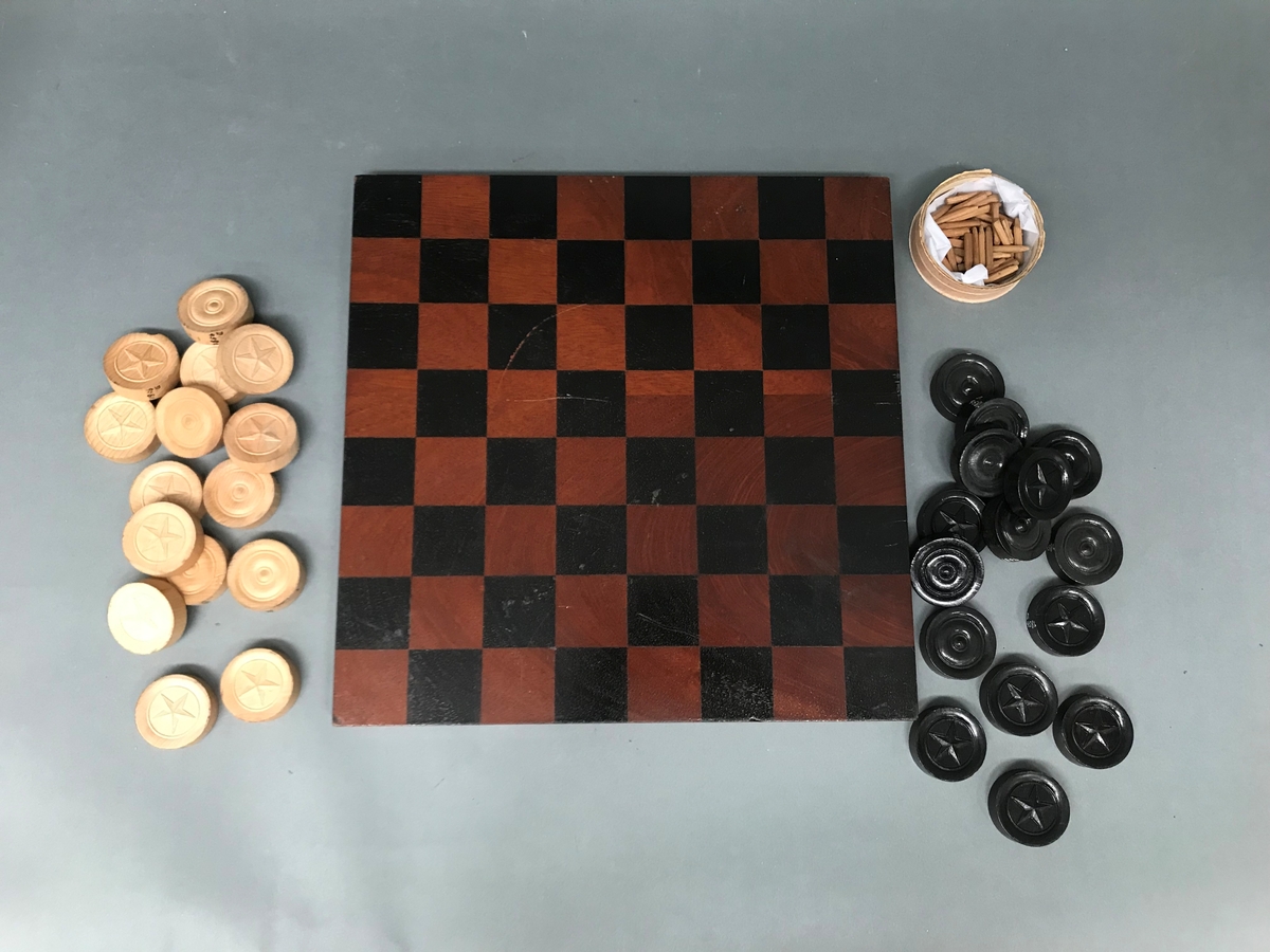 Brikker til brettspill. 15 hvite og 15 sorte dam-brikker og en eske med 45 små, runde pinner som smalner av nedover. To av pinnene er spadeformet. Brett av en kvadratisk, tykk mahognyplate med ruter på den ene siden (Dam) og korsformet rutesystem med diagonaler og huller i hjørnene på den andre siden (Rev og gjess-spill)