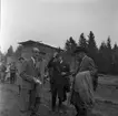 Skogsdag Örserums brunn 1961. Framför en brädgård står några män. De tittar sig omkring. Vissa av dem bär hatt. En man håller i en käpp. Skogen skymtar i bakgrunden.