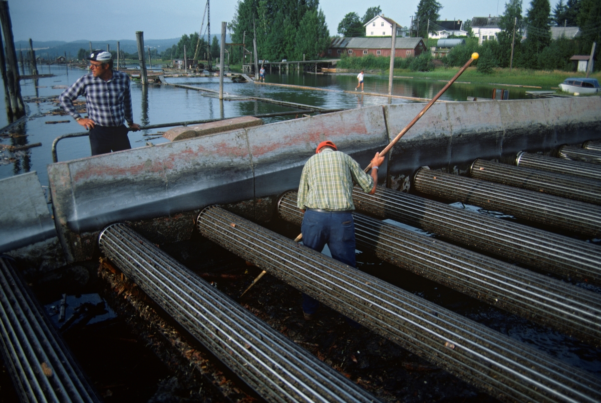 Fra retteanlegget ved Fetsund lenser i Akershus. De første to skrensrettingsmaskinene av denne nye typen installerte Glomma fellesfløtingsforening ved Glennetangen lenser i 1962-63. En av dem – den avbildete – ble flyttet til Fetsund i 1969. Noe høyere oppe i motstrøms retning (bak fotografen) var det en serie roterende piggvalser som tømmeret fra den ovenforliggene beholdningslensa skulle under. Hver valse hadde sin egen motor. Her ble tømmeret drevet i suksessivt økende hastighet fram mot ei «bru» – den konstruksjonen vi ser i den sentrale delen av dette fotografiet. Derfra ble virket kastet ned i en kum, som på dette bildet ligger skjult bak brua. Like ovenfor kummen var det et retterhjul på tvers av strømretningen som bidro til at stokkene kom rett og fint inn i renna. Her var det også et elektrisk drevet vannhjul som forsterket strømmen, og dermed bidro til at prosessen gikk noenlunde raskt. Da dette fotografiet ble tatt ser det ut til at man hadde stanset maskinen, antakelig for å fri den for fragmenter fra stokkene og annet rusk som hadde falt ned på den mekanikken som befant seg under brua. Bildet er antakelig fra 1984 eller 1985, en av de to siste sesongene Glomma ble brukt som transportåre for fløtingsvirke.