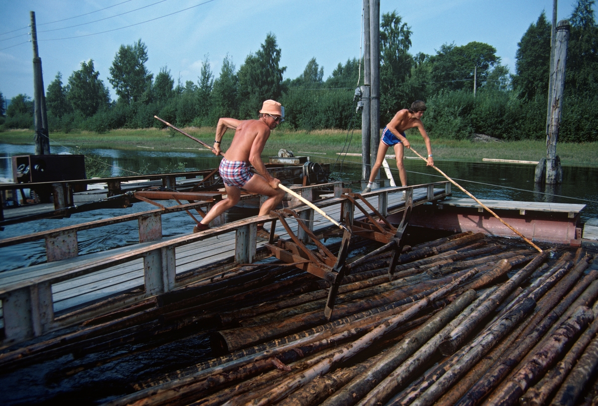 Asbjørn Østby fra skogbygda Setskog i Akershus sammen med en lokal skoleungdom i arbeid ved Fetsund lenser sommeren 1984. Begge var iført badeshorts og arbeidet med fløterhaker. Da dette fotografiet ble tatt sto karene antakelig på ei av bruene ved den såkalte «overkjøringa», der tømmeret (fra 1969 og framover) ble fordelt på rennene som skulle lede det videre mot de to mosemaskinene som fortsatt var i drift. Dette var et stressende arbeid, for mannskapene som betjente soppemaskinene skulle ikke behøve å vente for mye på tømmer til neste «soppe». Derfor ble tømmeret penset fra den ene til den andre renna med cirka et kvarters mellomrom. Asbjørn og arbeidskameratene hans måtte derfor finne seg i en del bebreidelser. I Thomas Støvind Bergs bok «Stemmer fra elva – Fløtingas historie i Fet» (2012) er Bjørn Holmedal sitert slik:

«Je huser at je var hærm mange ganger, når dem ikke greide å sende nok tømmer i begge løypa nerover tel inner- og yttermaskin’. Iblant fekk vi jo masse, men så kunne det bli blankt vann lenge nerover i renna vår og morsatt. Je husser at je likte det itte når innermaskin’ gikk for full fartog vi sto der og itte fekk kryssa ta no’. ‘Ja, je var litt hærm på’n Asbjørn iblant (latter). Jeg huser je sa ein gong tel’n «du kan itte kjøre så lenge på ein maskin!» Men du veit, detta visste jo han mye beire enn meg!»

Sjøl kommenterte Asbjørn Østby dette problemet slik:

«Gjorde du itte rekti på overkjøringa, måtte dom stoppe opp der oppe på maskin’. Men je kjørde jamt der oppe på overkjøringa, og heldt dom med tømmer, sa da var’e jo opp tel dom hvem som klarde å produsere mest.»

Hastigheten i tilførselen av tømmeret til mosemaskinene var nok også avhengig av strømforhold og vind, noe som i den siste driftsperioden ble forsøkt overvunnet ved hjelp av vaiertrekk.