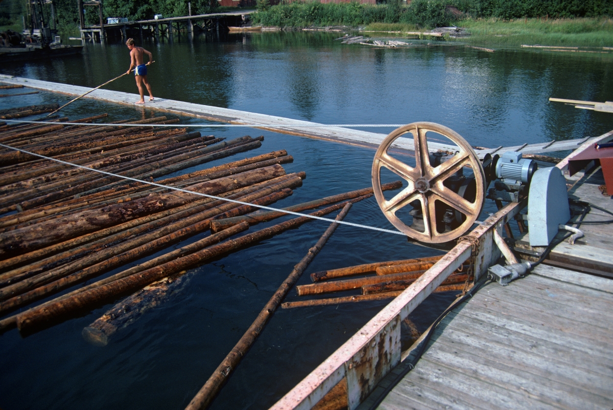 Vendepunktet for et av vaiertrekkene som skulle sørge for at tømmeret hadde god framdrift i ei av «rennene» ned mot ei av «mosemaskinene» ved Fetsund lenser. Disse rennene var 5-6 meter brede kanaler, avgrenset av flåteganger. Her var det meninga at tømmer som nettopp hadde passert retteanlegget og følgelig fløt parallelt, på tvers av rennas lengderetning, nedover mot den delen av anlegget der stokkene ble buntet. Lenseanlegget var plassert i et stilleflytende parti, litt ovenfor deltaområdet der hvor Glomma renner ut i innsjøen Øyeren. Stundom – og særlig når det var sønnavind – drev tømmeret svært langsomt nedover rennene, og dermed ble ekspedisjonshastigheten ved mosemaskinene langsom. Det var dette problemet vaiertrekket vi ser på fotografiet skulle bøte på. Trekkvaieren var endeløs – den roterte rundt hjul i driftsområdets ytterender. Hjulet som drev dette systemet ble satt i rotasjon av en elektromotor som ses på dette bildet. Den delen av vaiersløyfa som gikk underst berørte tømmerstokkene renna og pakket dem tett sammen i ett enkelt lag. Ved lenseanlegget på Glennetangen skulle vaieren gå med en hastighet på 18 centimeter i sekundet, og det er nærliggende å anta at framdriften her på Fetsund var omtrent den samme. Glennetangen fikk denne teknologien i 1962-63, på Fetsund kom den i 1969. Systemet innebar at det ikke lenger var behov for like mange som gikk på rennenes flåteganger med fløterhaker (jfr. gutten i bakgrunnen), der de rettet på og dro i stokkene som skulle ned mot mosemaskinene.