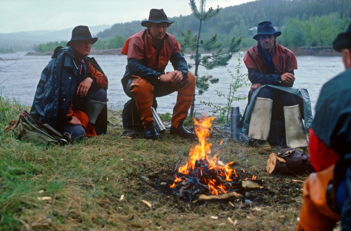 Fire fløtere, samlet rundt et bål ved Trysilelva, like sør for Jordet i Trysil, Hedmark. Fotografiet ble tatt en regnværsdag i 1987. Fløterne var iført fargerikt regntøy (jakker og bukser) og hadde vidbremmete filthatter på hodet. De hadde også ryggsekker – antakelig for nistematen – og ståltermoser med kaffe.