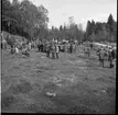 En folksamling står på en gräsmatta. En traktor står i mitten av folksamlingen. I bakgrunden finns en skog.