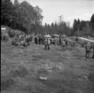 En folksamling står på en gräsmatta. En traktor står i mitten av folksamlingen. I bakgrunden finns en skog.