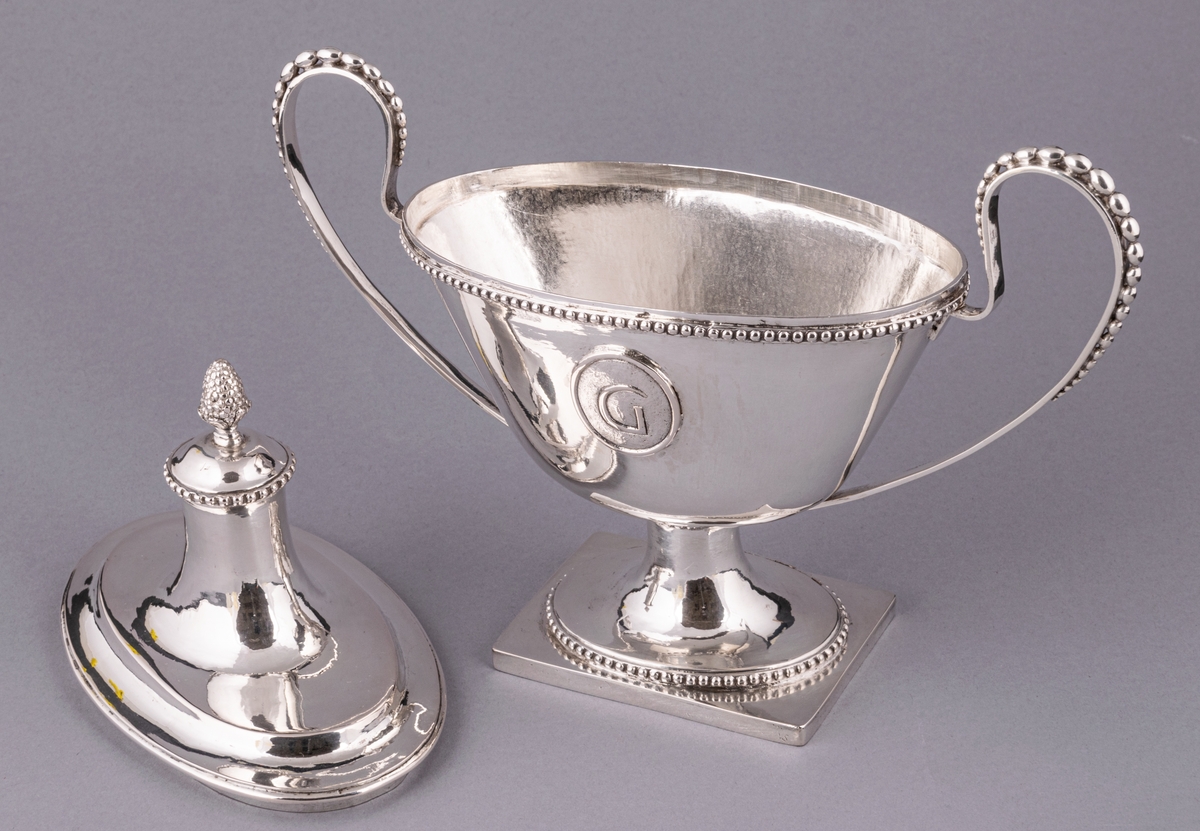 En av ett par sengustavianska sockerskålar i silver stämplade Daniel Elfbom, Gävle 1791. Oval skål på rektangulär fot. Löst lock med knopp och två utvändiga uppsvängda hänklar med pärlstav. Pålödd medaljong med Gävle stads vapen (G). Stämpar på fotens kant: DE G I 2 och tre kronor.