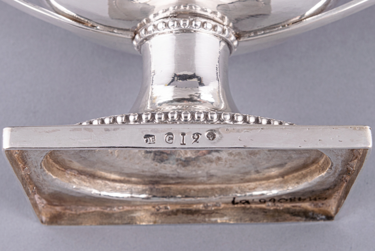 En av ett par sengustavianska sockerskålar i silver stämplade Daniel Elfbom, Gävle 1791. Oval skål på rektangulär fot. Löst lock med knopp och två utvändiga uppsvängda hänklar med pärlstav. Pålödd medaljong med Gävle stads vapen (G). Stämpar på fotens kant: DE G I 2 och tre kronor.