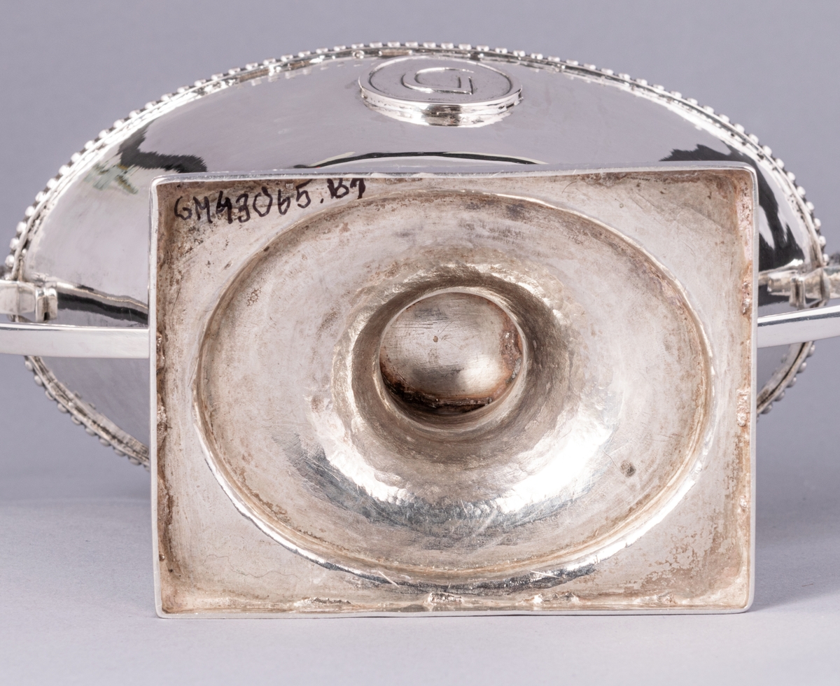 En av ett par sengustavianska sockerskålar i silver stämplade Daniel Elfbom, Gävle 1791. Oval skål på rektangulär fot. Löst lock med knopp och två utvändiga uppsvängda hänklar med pärlstav. Pålödd medaljong med Gävle stads vapen (G). Stämpar på fotens kant: DE G I 2 och tre kronor.