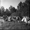 Några ungdomar och barn står uppställda för fotografering. De tittar på en man som står och gestikulerar. I bakgrunden syns uppslagna tält.