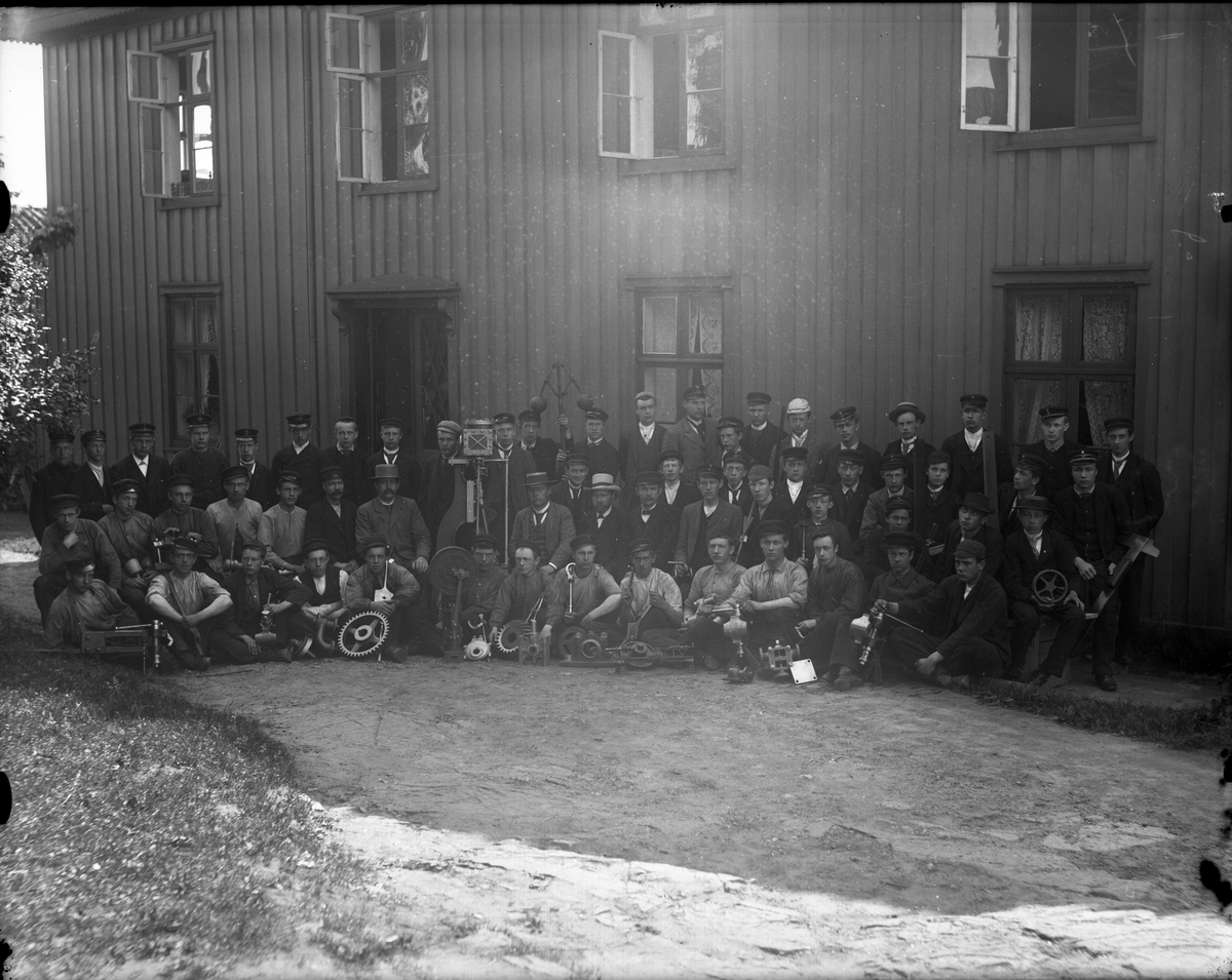 Fotosamling etter fotograf John Nielsen, Porsgrunn. Gruppeportrett fra Skiensfjorden Mekaniske Fagskole på Osebakken. Elevene har med seg gjenstander som beskriver skolens innhold. Bildet er tatt foran Rasmus Jørgensen Brønlund sitt hus. Brønlund var skolens stiften og første Rektor. Huset lå på vestsiden ved Porsgrunn Verksted.