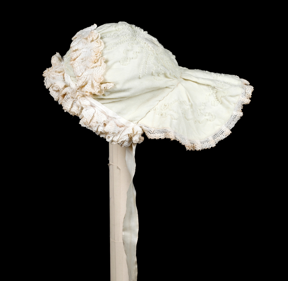 Kråke i hvit, tynn ull med ullbroderier på både pull og nakkestykke. Broderiet består av små løkker i bølgende mønster. Innfelt silkestykke på bakhodet og kanting av blonde, sannsynligvis av bomull. Luen er fôret med fint bomullslerret. Knytebånd i sateng, muligens silke.