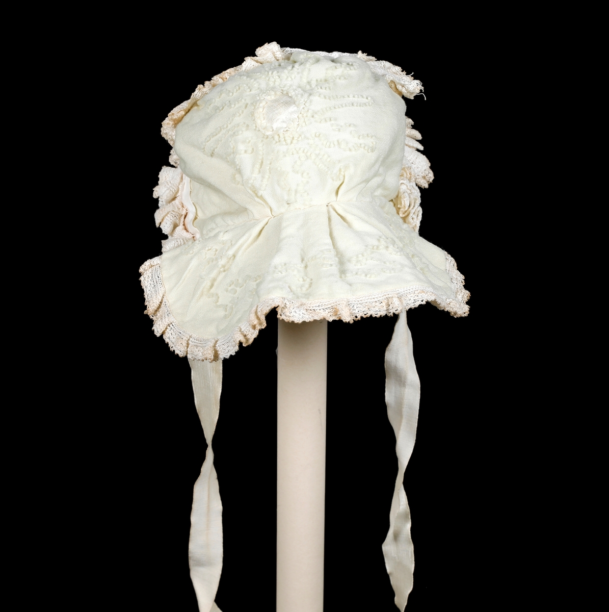 Kråke i hvit, tynn ull med ullbroderier på både pull og nakkestykke. Broderiet består av små løkker i bølgende mønster. Innfelt silkestykke på bakhodet og kanting av blonde, sannsynligvis av bomull. Luen er fôret med fint bomullslerret. Knytebånd i sateng, muligens silke.