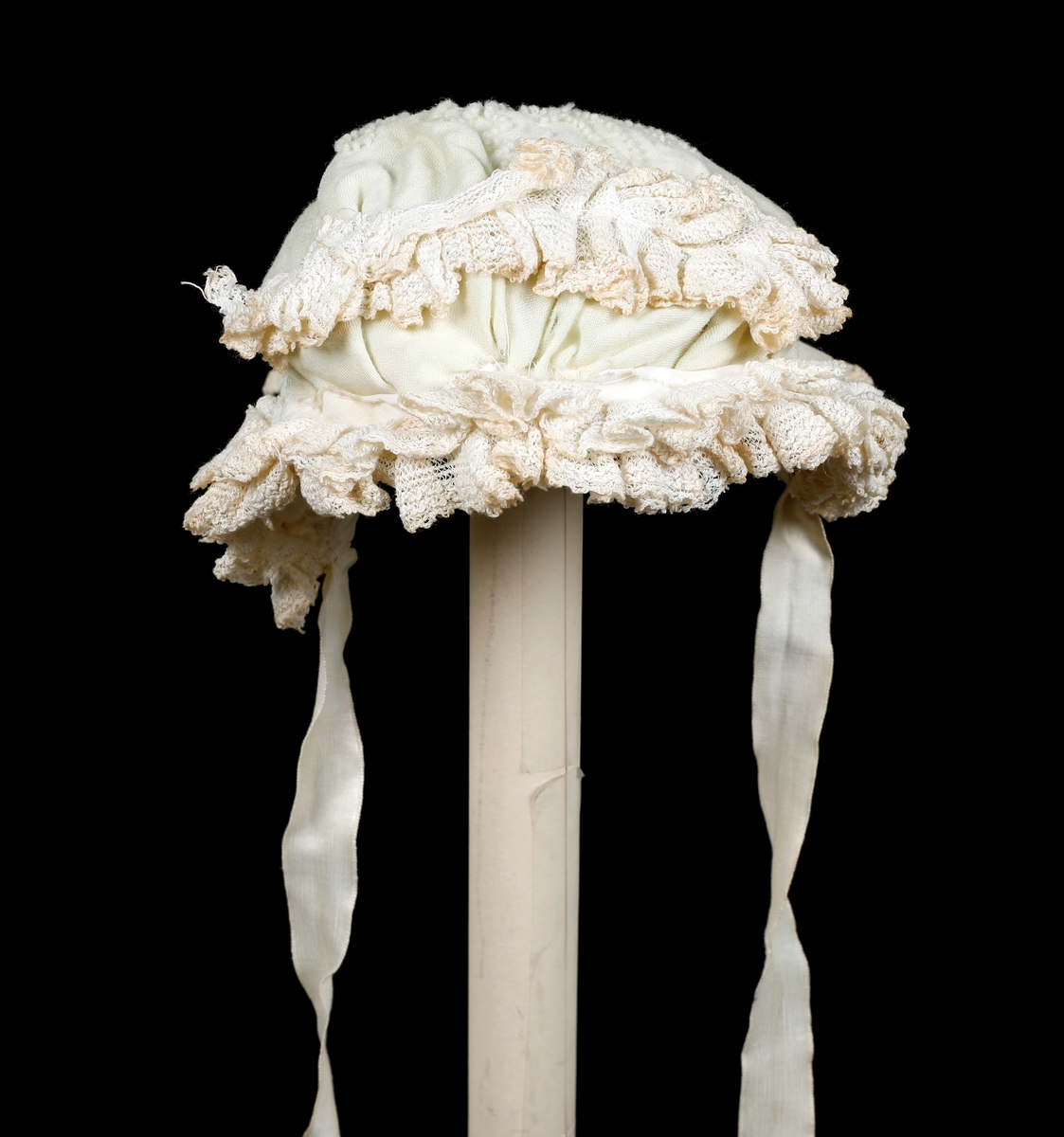 Kråke i hvit, tynn ull med ullbroderier på både pull og nakkestykke. Broderiet består av små løkker i bølgende mønster. Innfelt silkestykke på bakhodet og kanting av blonde, sannsynligvis av bomull. Luen er fôret med fint bomullslerret. Knytebånd i sateng, muligens silke.