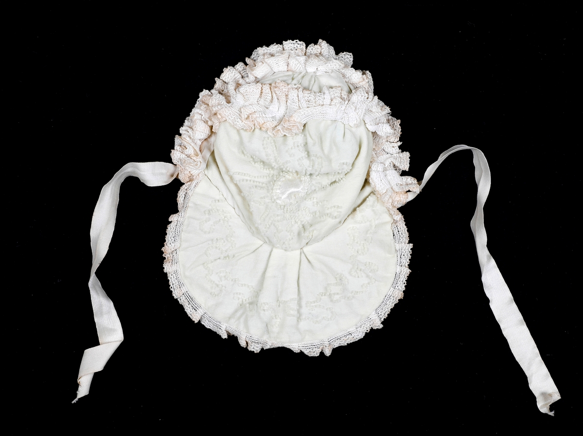 Kråke i hvit, tynn ull med ullbroderier på både pull og nakkestykke. Broderiet består av små løkker i bølgende mønster. Innfelt silkestykke på bakhodet og kanting av blonde, sannsynligvis av bomull. Luen er fôret med fint bomullslerret. Knytebånd i sateng, muligens silke.