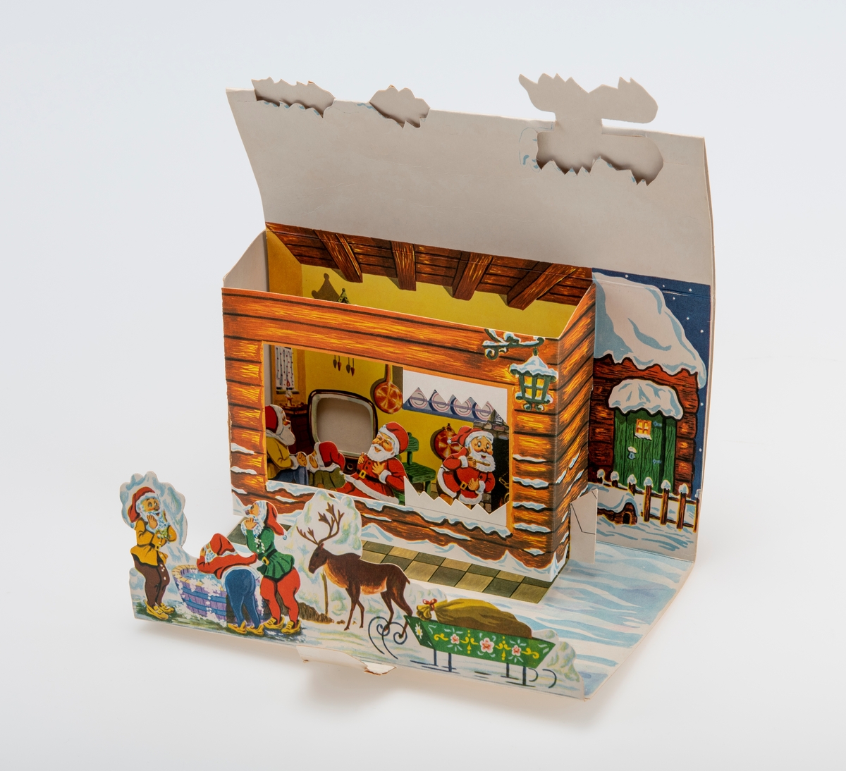 En adventskalender laget av kartong. Sammenbrettet er den flat. På den ene siden er det felt for frimerke/porto og for å skrive på navn/adresse for å kunne sende den i posten. På en andre siden er det påtrykt motiv som viser en husvegg med en grønn inngangsdør og et vindu som er opplyst. Mot veggen står det en trillebår, en bøtte, en benk med en stamp på og en stige. Dette utgjør baksiden på kalenderen når den er oppslått. Når kalenderen er oppslått er den tredimensjonal. I front brettes opp kartong som viser tre nisser rundt en vaskebalje og et reinsdyr med slede. på sleden er det en gavesekk. Deretter er det et felt med "sne". Der er kalenderlukene 1-8. Deretter er det en "tømmerbygning" som stikker ut fra bakgrunnen. I et "vindu" kan man se fire nisser i et kombinert kjøkken/stue. Til høyre er det en grue og på bakveggen er det en tallerkenhylle og et gjøkur. I venstre hjørne står det en tv/et fjernsyn. På høyre side av kalenderen er det en åpning med en pappremse med illustrasjoner. Pappremsen har ulike illustrasjoner som utgjør TV-bilder på nissenes fjernsyn. Kalenderluke 9-24 er på taket. Når man åpner kalenderlukene er det ulike tegninger.
Ved kalenderen lå det ved to pappfigurer. Den ene viser en nisse som står og inn i et vindu som viser en stue med pyntet juletre. Den andre figuren viser en nisse som står ved det snødekt tre. Midten av treet er skåret ut slik at det utgjør en ramme. Det er usikkert om disse originalt tilhører kalenderen.