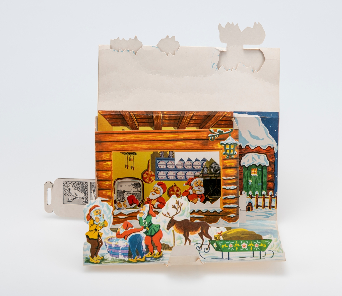 En adventskalender laget av kartong. Sammenbrettet er den flat. På den ene siden er det felt for frimerke/porto og for å skrive på navn/adresse for å kunne sende den i posten. På en andre siden er det påtrykt motiv som viser en husvegg med en grønn inngangsdør og et vindu som er opplyst. Mot veggen står det en trillebår, en bøtte, en benk med en stamp på og en stige. Dette utgjør baksiden på kalenderen når den er oppslått. Når kalenderen er oppslått er den tredimensjonal. I front brettes opp kartong som viser tre nisser rundt en vaskebalje og et reinsdyr med slede. på sleden er det en gavesekk. Deretter er det et felt med "sne". Der er kalenderlukene 1-8. Deretter er det en "tømmerbygning" som stikker ut fra bakgrunnen. I et "vindu" kan man se fire nisser i et kombinert kjøkken/stue. Til høyre er det en grue og på bakveggen er det en tallerkenhylle og et gjøkur. I venstre hjørne står det en tv/et fjernsyn. På høyre side av kalenderen er det en åpning med en pappremse med illustrasjoner. Pappremsen har ulike illustrasjoner som utgjør TV-bilder på nissenes fjernsyn. Kalenderluke 9-24 er på taket. Når man åpner kalenderlukene er det ulike tegninger.
Ved kalenderen lå det ved to pappfigurer. Den ene viser en nisse som står og inn i et vindu som viser en stue med pyntet juletre. Den andre figuren viser en nisse som står ved det snødekt tre. Midten av treet er skåret ut slik at det utgjør en ramme. Det er usikkert om disse originalt tilhører kalenderen.