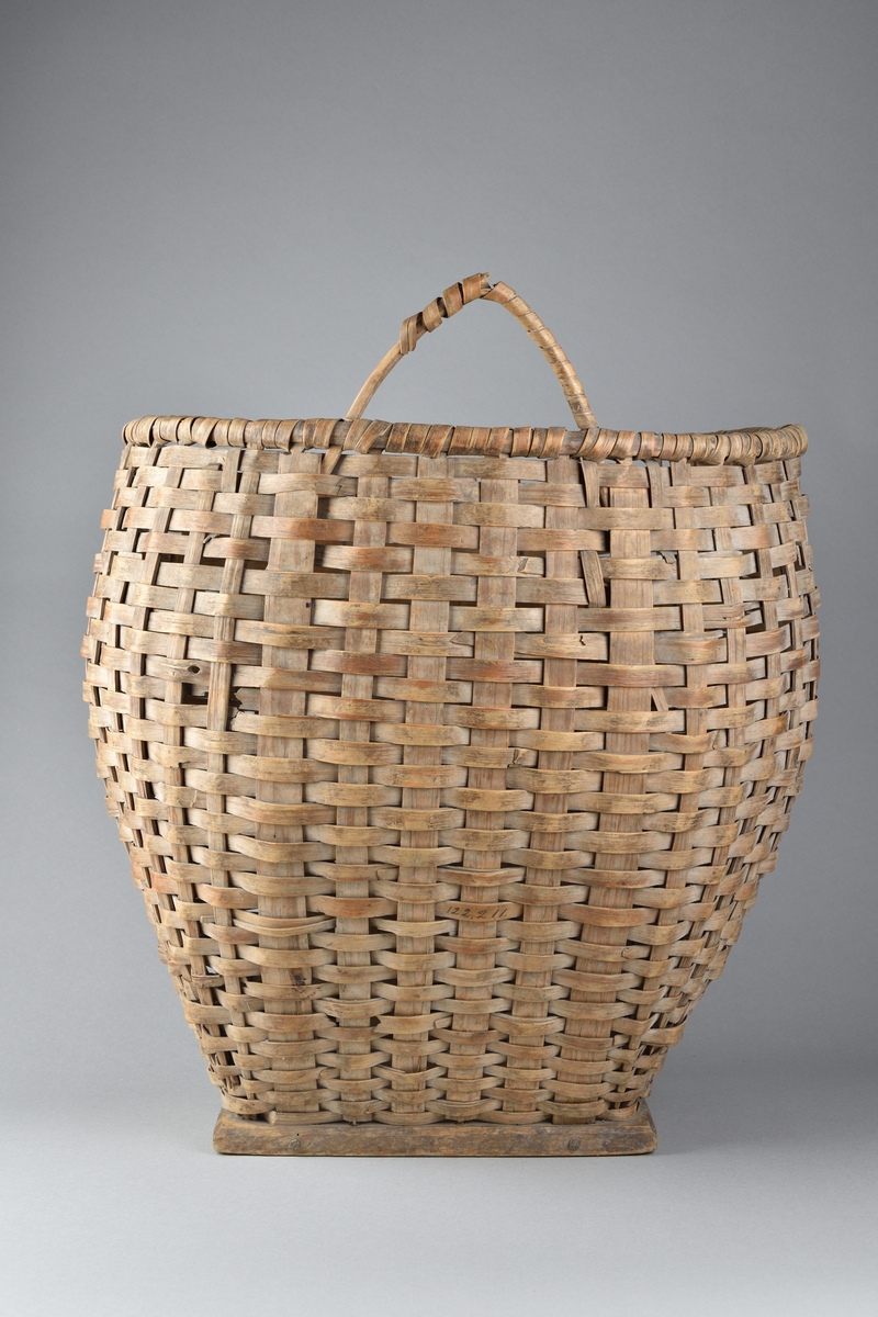 Ullkorg tillverkad av flätat träspån. Rund och bukig överdel, med bred öppning samt med ett handtag. Nederdelen avsmalnande. Korgen vilar på en rektangulär fotplatta av trä.
Korgen har möjligtvis haft ett ytterligare handtag vilket numera saknas. 