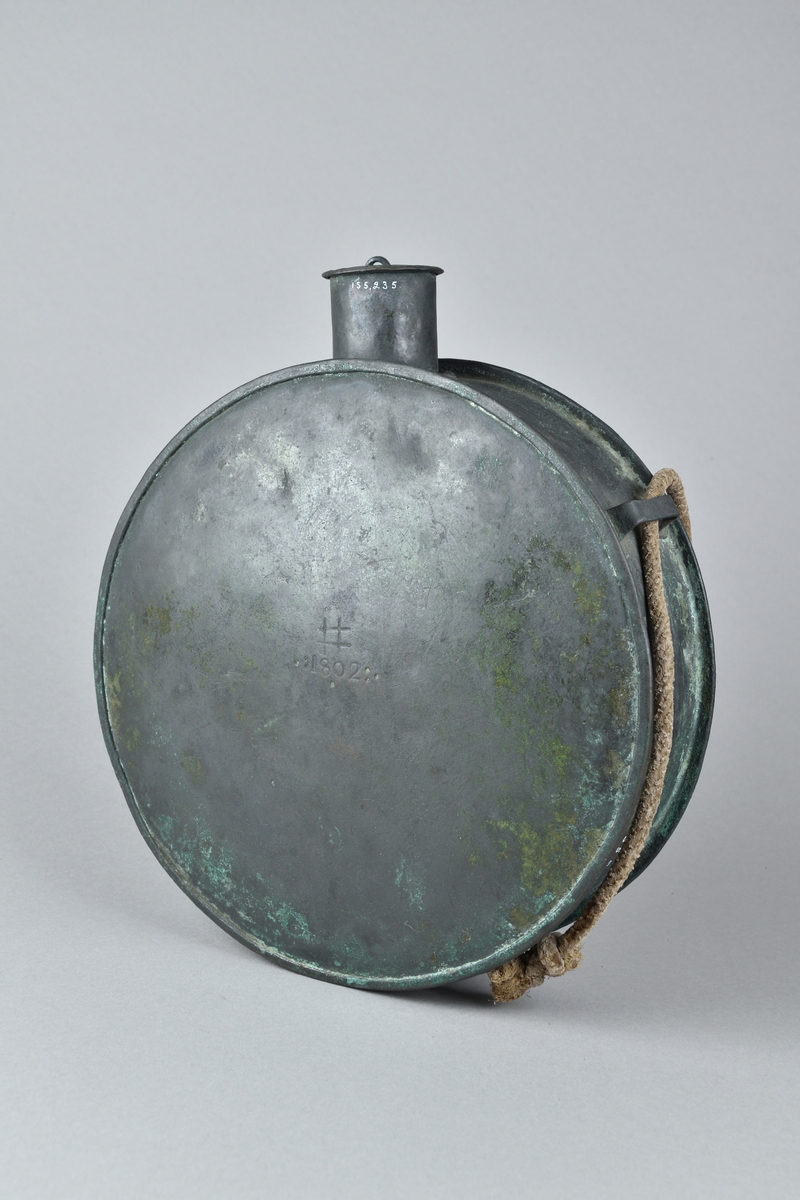 Krutflaska tillverkad i koppar. Stor, rund och platt, med kort hals och lock. Ett snöre för upphängning runt flaskan, hållet på plats av tre fästen. Flaskans framsida med stämpel: "# 1802". 