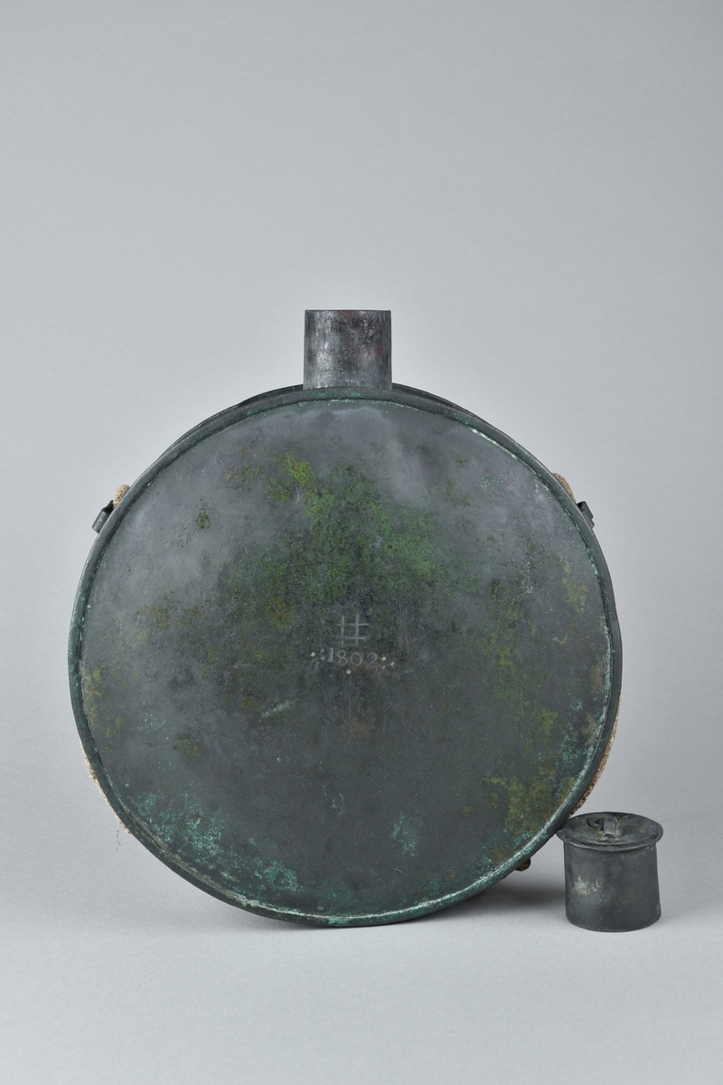 Krutflaska tillverkad i koppar. Stor, rund och platt, med kort hals och lock. Ett snöre för upphängning runt flaskan, hållet på plats av tre fästen. Flaskans framsida med stämpel: "# 1802". 