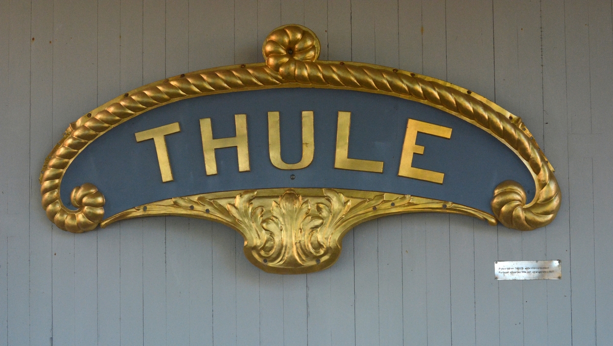 Akterstävsornament av mässing. I mitten av ornamentet står texten: "THULE". Kanten på ornamentets övre halva är täckt med ett förgylldt rep. Den undre delen visar tre förgyllda löv. Förutom den förgyllda färgen har ornamentet en grå-blå bakgrund.