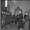 Några unga scouter står uppradade framför en scoutledare i en kyrka. Scoutledaren stå och håller tal.