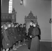 Några unga scouter står uppradade framför en scoutledare i en kyrka. Scoutledaren stå och håller tal.