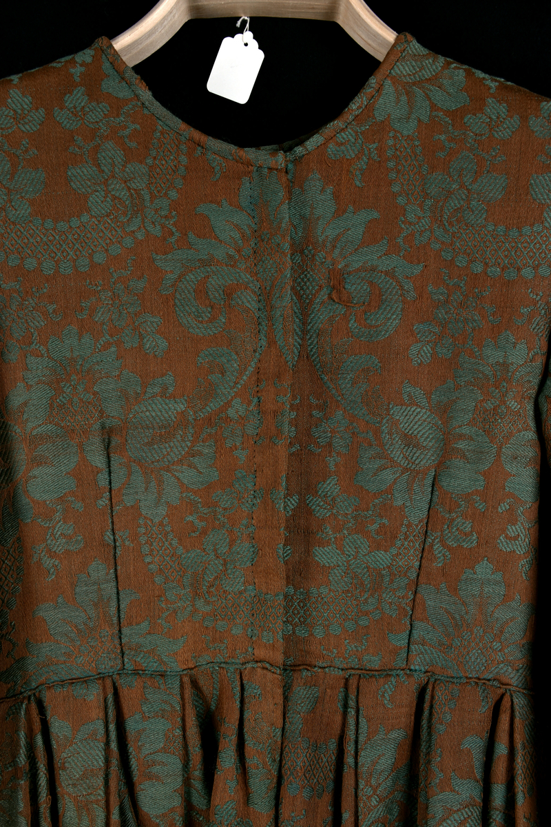 Handsydd bröllopsklänning i jacquardväv med grönt växtmönster mot brun bakgrund. Främre knäppning med nio hyskor, högknäppt och enkel halsring. Hellång ärm och kjol. Foder i ofärgat- respektive rutmönstrat linnetyg. Visst bruksslitage.