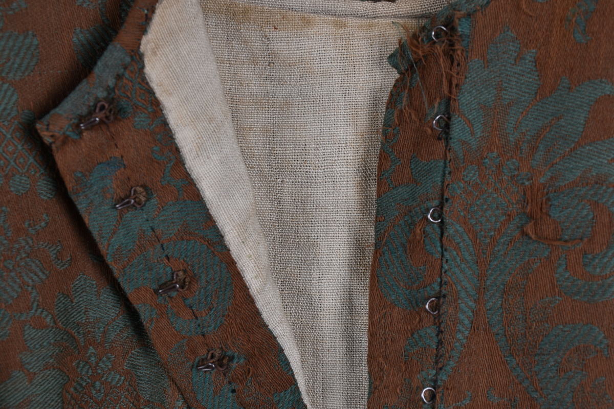 Handsydd bröllopsklänning i jacquardväv med grönt växtmönster mot brun bakgrund. Främre knäppning med nio hyskor, högknäppt och enkel halsring. Hellång ärm och kjol. Foder i ofärgat- respektive rutmönstrat linnetyg. Visst bruksslitage.