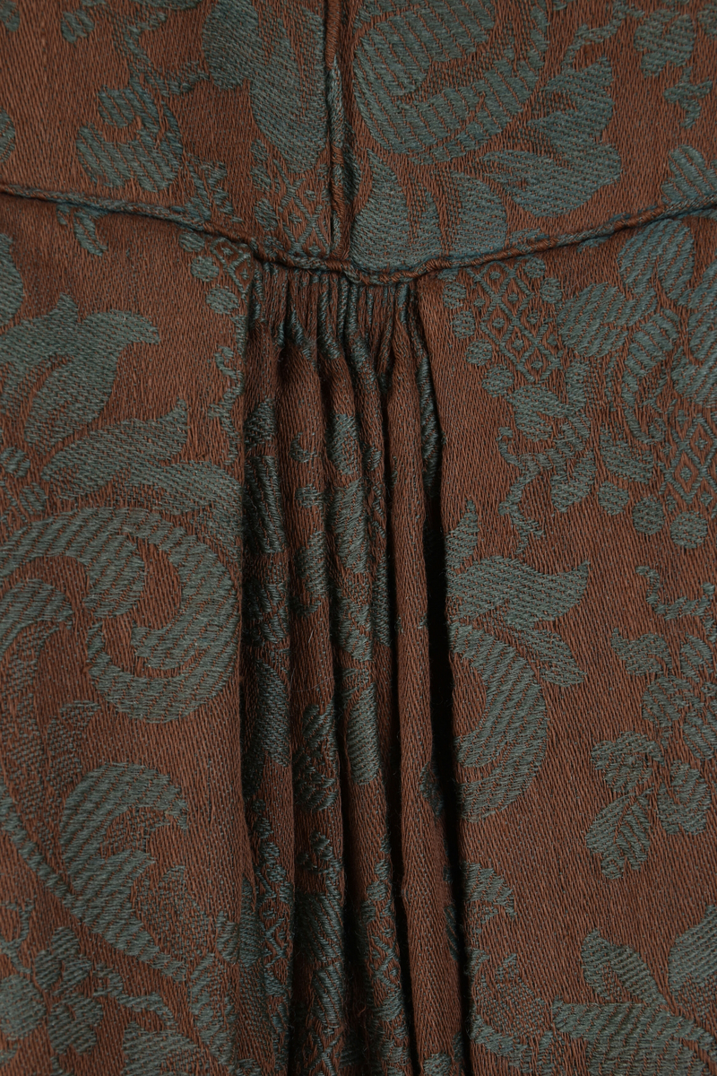 Handsydd bröllopsklänning i jacquardväv med grönt växtmönster mot brun bakgrund. Främre knäppning med nio hyskor, högknäppt och enkel halsring. Hellång ärm och kjol. Foder i ofärgat- respektive rutmönstrat linnetyg. Visst bruksslitage.