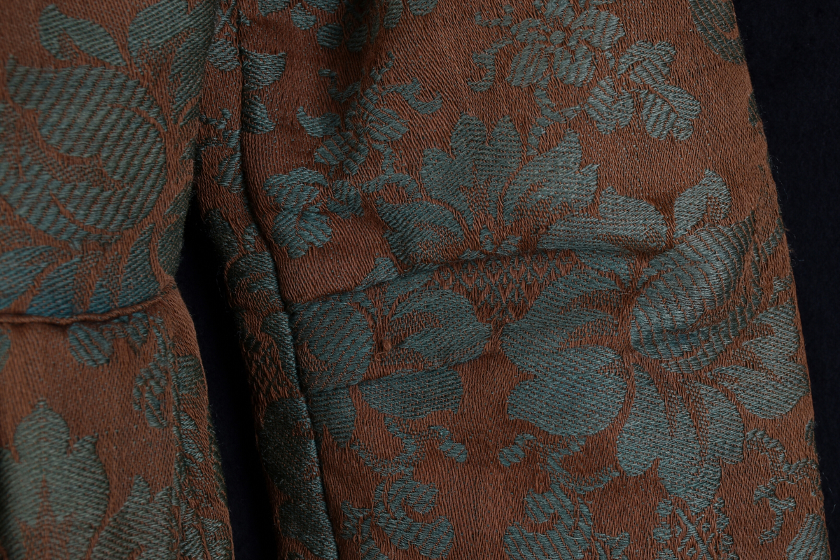 Handsydd bröllopsklänning i jacquardväv med grönt växtmönster mot brun bakgrund. Främre knäppning med nio hyskor, högknäppt och enkel halsring. Hellång ärm och kjol. Foder i ofärgat- respektive rutmönstrat linnetyg. Visst bruksslitage.